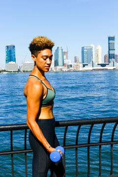 Athletic woman flexing with dumbbell by waterfront in New York City on sunny day