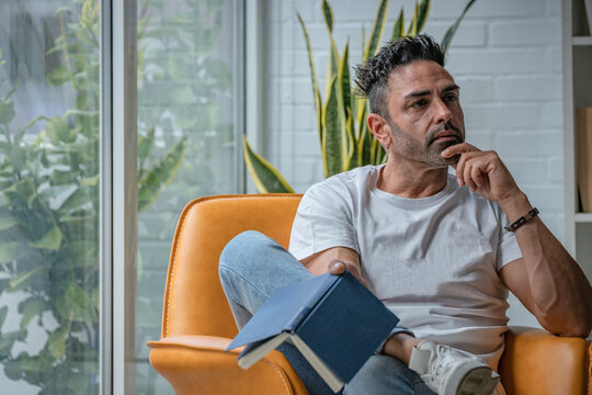 Middle-aged man in casual attire pausing from reading, gazing pensively into the distance while seated in a modern, light-filled living space