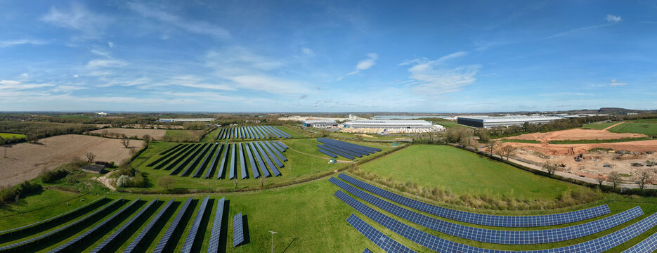 Aerial view of a large solar farm with rows of blue photovoltaic panels set in green fields near industrial warehouses under a clear blue sky in Coalville, England, United Kingdom.