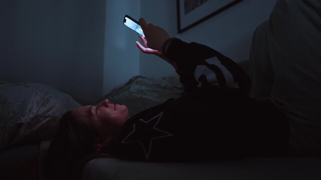 Girl doom scrolling her smartphone on a coach at night. Social network and phone addiction concept. Cozy home ambient light