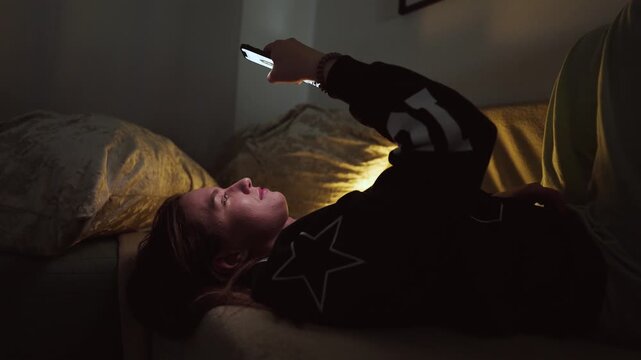 Girl doom scrolling her smartphone on a coach at night. Social network and phone addiction concept. Cozy home ambient light