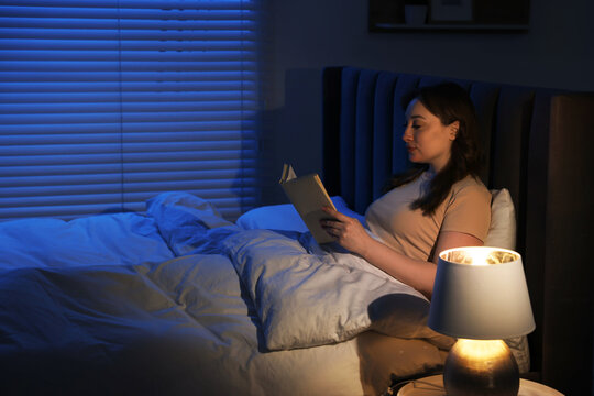 Woman reading book before sleep with night lamp in bed at home