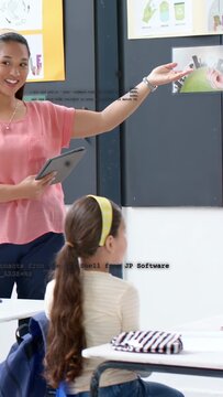 Vertical video: Teacher checking tablet and gesturing toward recycling poster, guiding class focus