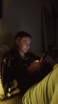 Girl doom scrolling her smartphone on a coach at night. Social network and phone addiction concept. Cozy home ambient light. vertical video