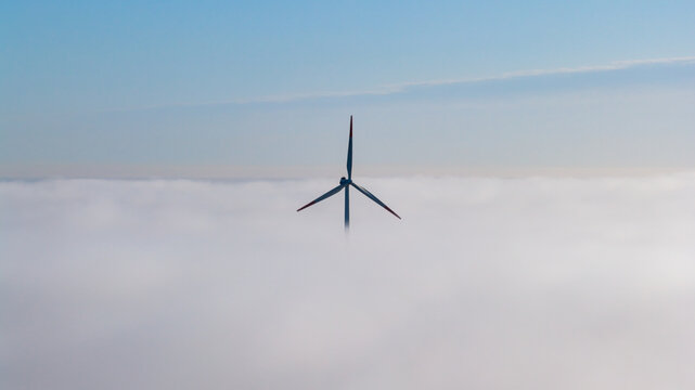 Aerial view of a single wind turbine with red-tipped blades emerging from a thick layer of white fog under a clear blue sky in Wislinka, Pomeranian Voivodeship, Poland.