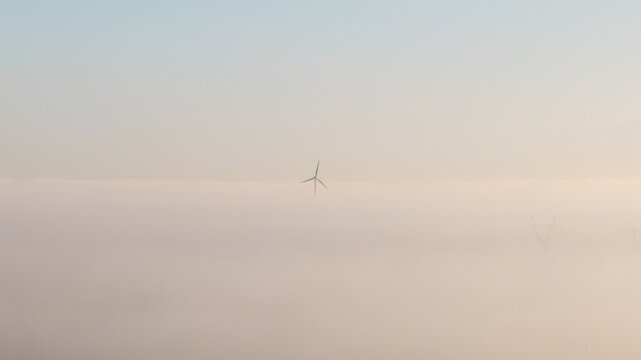 Aerial view of a single wind turbine rising above a thick layer of morning fog under a clear sky in Wislinka, Pomeranian Voivodeship, Poland.