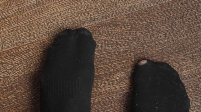 Top view of male feet in worn out black socks with a hole on the big toe. Person awkwardly shifting from foot to foot on a wooden floor