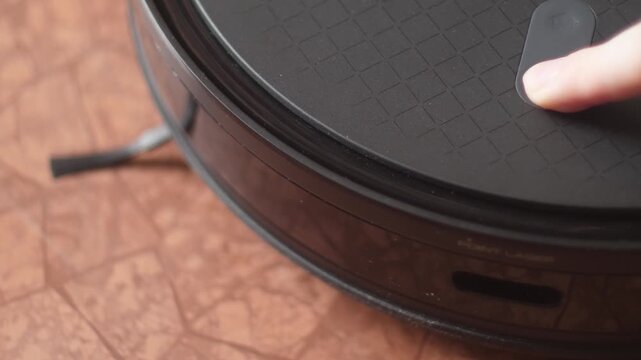 Person presses the power button on a robot vacuum cleaner to start cleaning. Modern smart gadget turns on with a light indicator on the floor