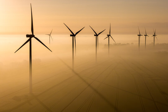 Aerial view of a wind farm with turbines standing in thick morning mist over agricultural fields during sunrise in Wislinka, Pomeranian Voivodeship, Poland.