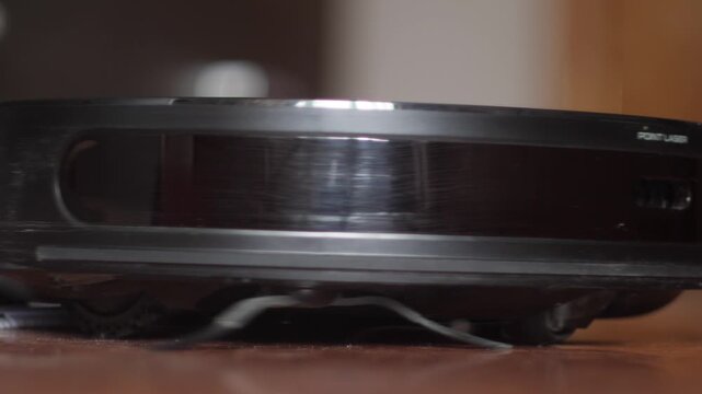 A black robot vacuum cleaner rotates in place and begins moving across a wooden floor. Side view of the sensors working and the automatic cleaner moving in the apartment