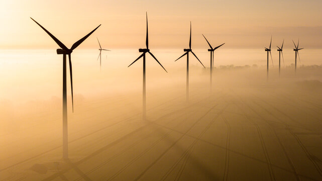 Aerial view of wind turbines standing in a foggy field during a golden sunrise with long shadows across the rural landscape in Wislinka, Pomeranian Voivodeship, Poland.
