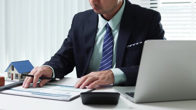 
Businessman shields tiny house with hand. Office desk holds coffee cup and pens. Professional demeanor suggests protection or security. Ideal for insurance, real estate, or financial services