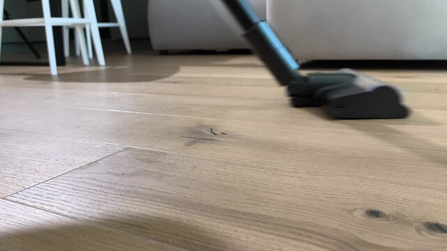 Vacuum cleaner removes dust from wooden floor in living room interior. Upright device cleans parquet surface using rotating brush system. Home cleaning routine maintains hygiene and order