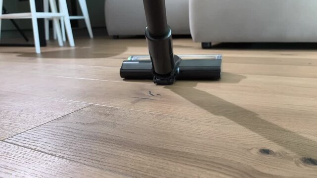 Vacuum cleaner brush moves over wooden floor removing dust particles. Close view shows rotating head cleaning parquet surface effectively. Home cleaning routine maintains neat interior