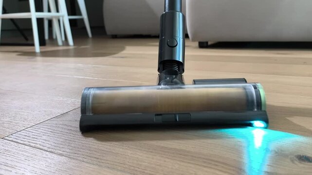 Vacuum cleaner brush moves toward camera cleaning wooden floor with light. Device head travels forward removing dust from surface. Home routine emphasizes efficient cleaning technology in