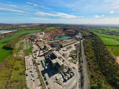 Aerial view of Cliffe Hill Quarry with industrial infrastructure, conveyor belts, and a green pond under a blue sky in Coalville, England, United Kingdom.