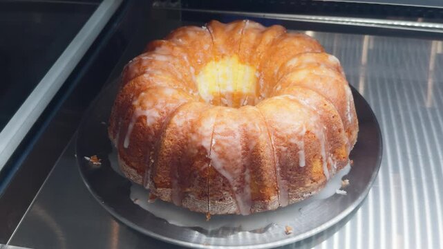 Golden bundt cake with sweet white glaze sitting on a silver platter, displaying several slices already cut, offering a tempting and delicious dessert option from a bakery display