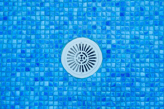 Overhead close-up of a swimming pool drain surrounded by blue mosaic tiles and clear water, highlighting symmetry, texture, and modern minimalist design
