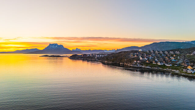 Aerial view of the coastal town of Nuuk with colorful houses along the rocky shoreline and calm water under a golden sunset sky with distant mountains Nuuk, Sermersooq, Greenland.