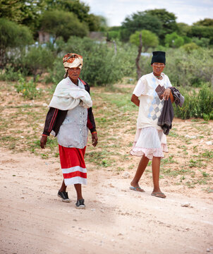 two Basarwa women walk through a sandy landscape with green bushes in Botswana
