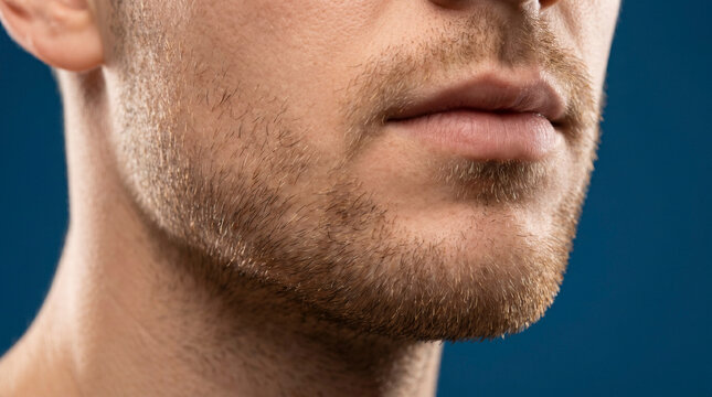 Macro photo of a man's lower face and jaw with thick stubble and mustache, lips slightly parted, detailed skin texture, over a solid dark blue background, side profile view.