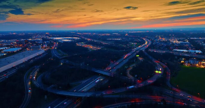 Vast scenery of the city against orange sky. Many cars with headlights on go by the roads, trestles and loops in the interchange. Aerial view.