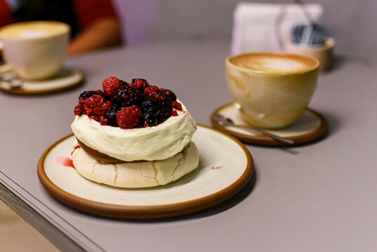 Elegante mini pavlova coronada con crema batida y frutos rojos frescos, servida con una taza de caf&eacute; en una mesa de cafeter&iacute;a. Fotograf&iacute;a con bokeh de postres gourmet para marketing culinario.