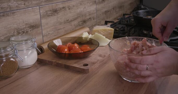 Female hands mixing raw minced meat with spices in a glass bowl using a spoon, preparing a homemade recipe on a wooden kitchen countertop with fresh ingredients nearby for a delicious dinner.