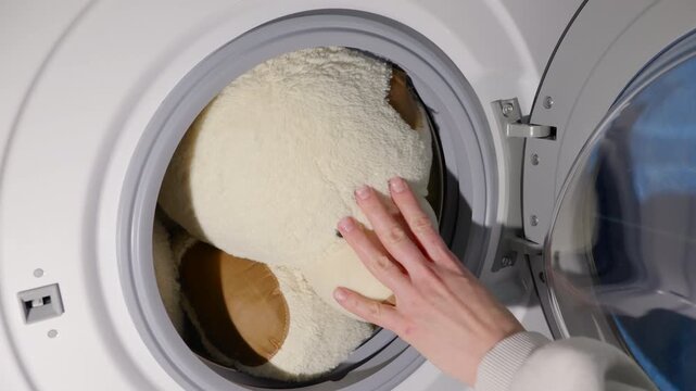 Washing a large plush teddy bear in a machine. Close-up view of a person's hand placing a large, fluffy teddy bear into a modern washing machine and closing the door