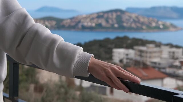 Person admiring a beautiful coastal town view from balcony. Close-up of a hand resting on a balcony railing while the focus shifts to a stunning view of a seaside town with islands and blue water