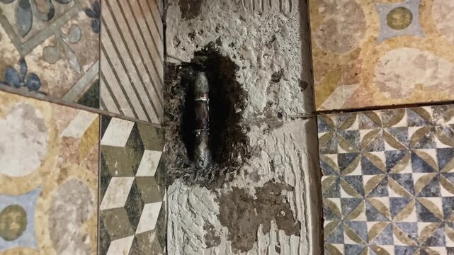 Slow push in toward damaged mosaic tile wall revealing exposed copper pipe and repaired plumbing inside broken concrete section