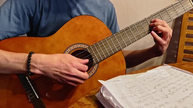 Close up of young man playing acoustic guitar with handwritten music notes on table during practice session indoors