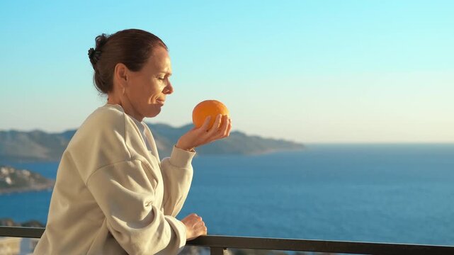 Woman with loss of smell trying to sense an orange. Mature woman on a balcony with a beautiful sea view experiencing anosmia or hyposmia, attempting to smell a fresh orange but feeling frustrated