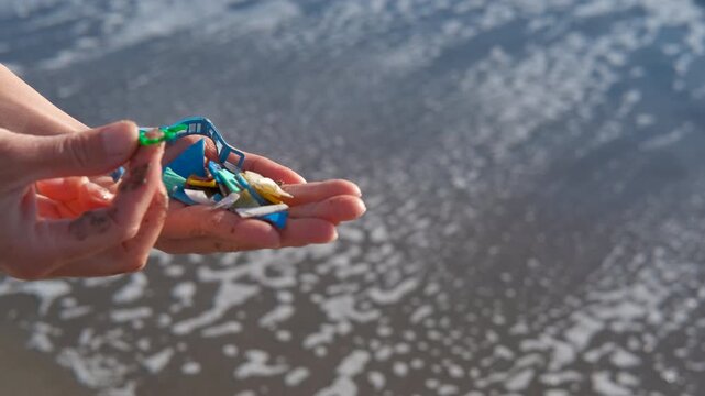 Person holding microplastics collected from the ocean shore. Close-up of hands holding many colorful pieces of microplastic debris collected from the beach, with the gentle sea waves