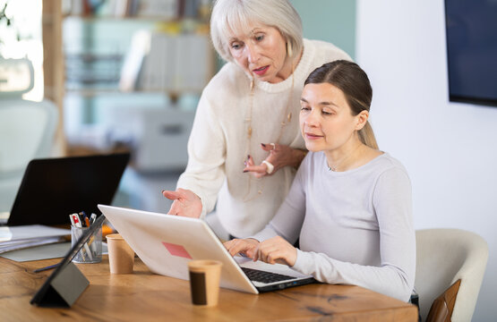Elderly man helps young female colleague use laptop while working in office space