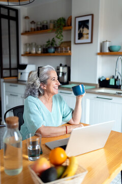 Senior woman enjoying coffee at home kitchen with laptop