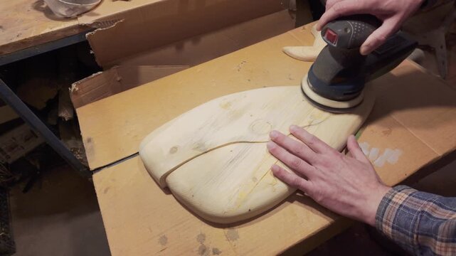 A artisan&rsquo;s hand guides an orbital sander over the surface of a handcrafted wooden whale sculpture. This close-up view focuses on the natural wood grain and the repetitive, tactile nature of the sandi