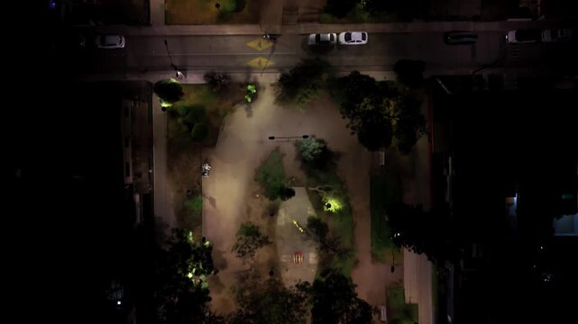 Aerial top-down night view of a public square from 75 meters height, street lights illuminating trees, gravel paths and human silhouettes in Plaza de Armas, 4K cinematic drone stock footage.