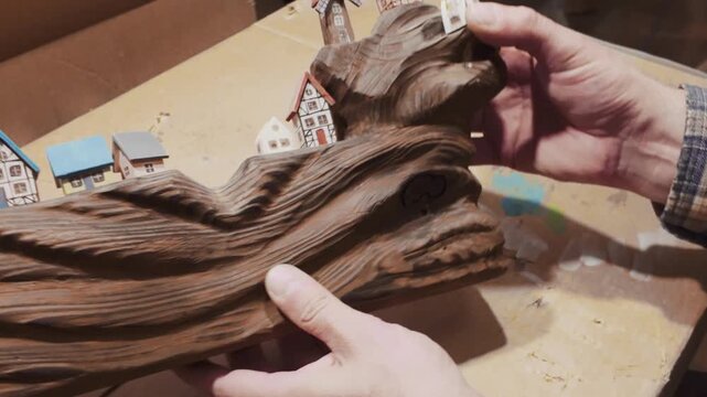 A close-up shot of hands carefully arranging miniature wooden houses and a windmill on a beautifully textured, dark wood driftwood base.