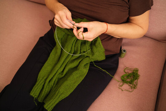 Person knits a green sweater in a cozy indoor space 