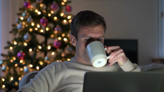 Man using laptop while sitting on sofa near Christmas tree at home to work or study. Casual man relaxing with computer and drinking coffee in home interior decorated for winter holidays