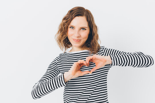 Heart gesture made by smiling woman in striped top against white backdrop