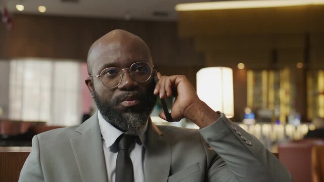 Professional Black businessman with beard and glasses, dressed in suit and tie, talking on mobile phone in lobby of upscale hotel