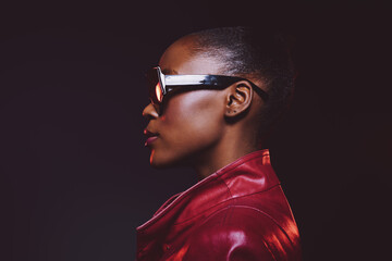 Profile portrait of a confident black woman with dark skin wearing stylish glasses and a red leather jacket against a dark background. Concept for fashion, empowerment, and modern style. © SHOTPRIME STUDIO
