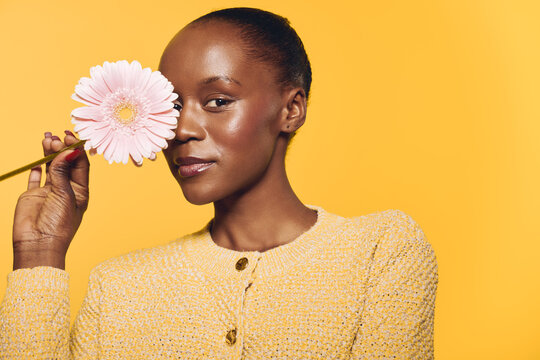 Portrait of a young black woman with dark skin holding a pink flower near her face. The yellow background and cozy sweater create a warm and cheerful mood. Concept of beauty and nature.
