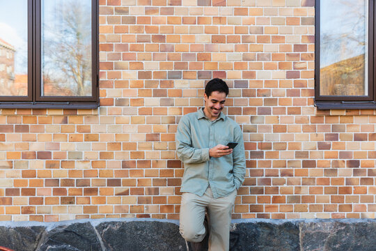 Man Leaning Against Brick Wall Using Smartphone Outdoors