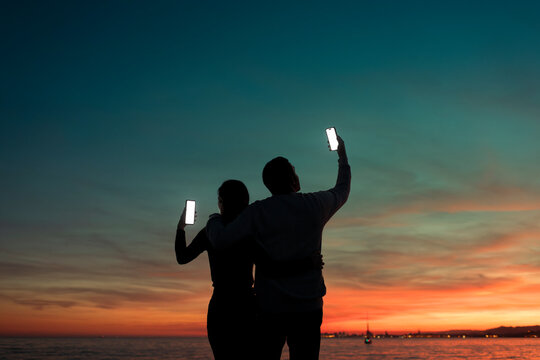 Hugged couple enjoying scenic sunset through phones