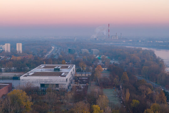 Aerial view of the modern Polish History Museum building surrounded by autumn trees near the Vistula river under a pink sunset sky in Warsaw, Masovian Voivodeship, Poland.