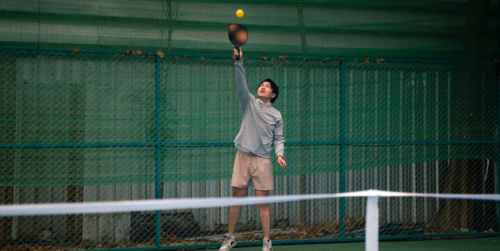 Young man playing pickleball on court, reaching up to hit yellow ball