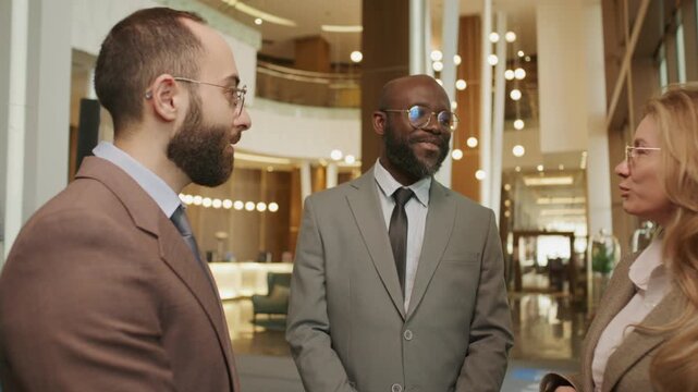 Team of three diverse professionals in formalwear standing together in upscale hotel lobby and discussing business while traveling for work
